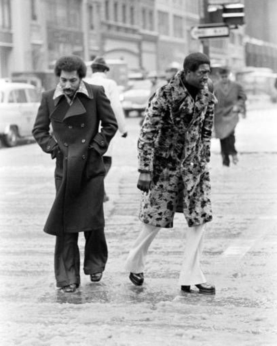 Two men walking through a snowy city street wearing long vintage coats and flared trousers.