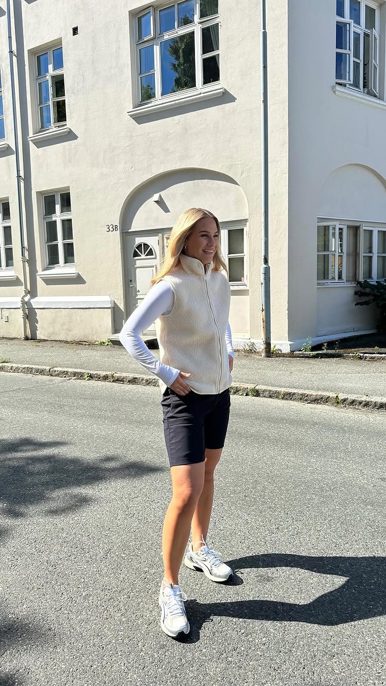 Woman wearing a white fleece vest over a long-sleeved shirt with black shorts and white sneakers.