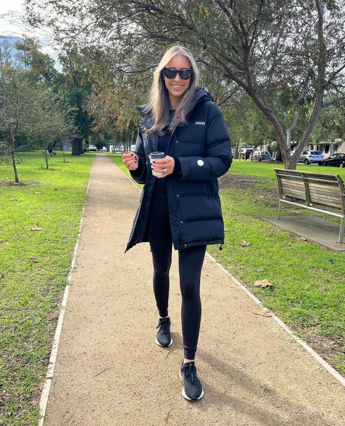 Blonde woman in a black puffer jacket and leggings walking through a park with coffee.