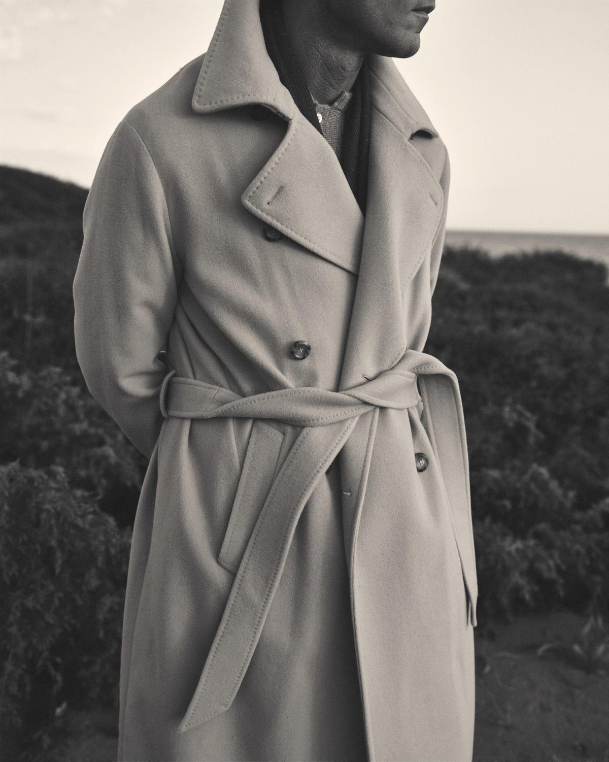 A person wearing a long, belted cashmere coat standing against a natural landscape background.