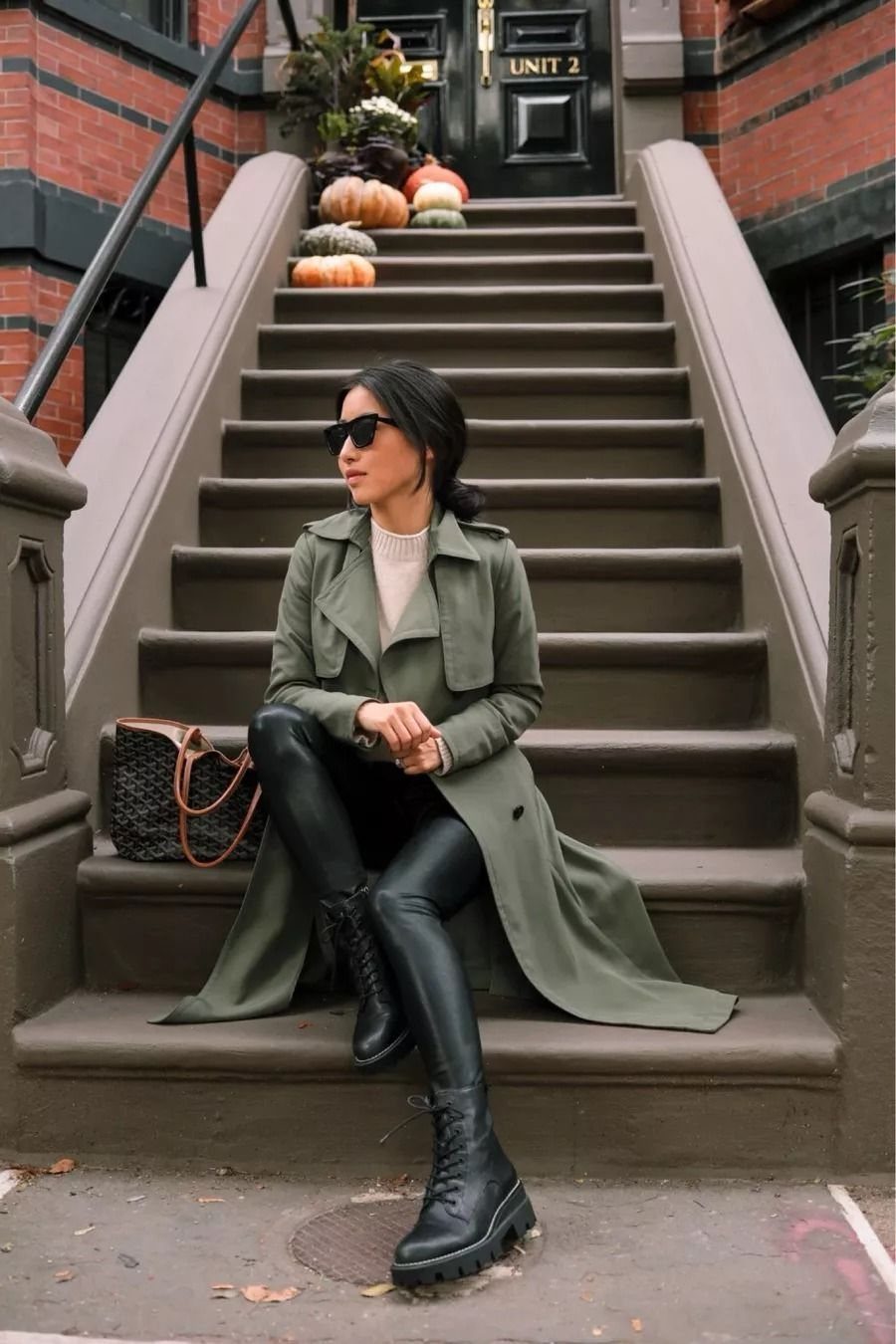 Woman in an olive trench coat and black leather leggings sitting on outdoor stairs.