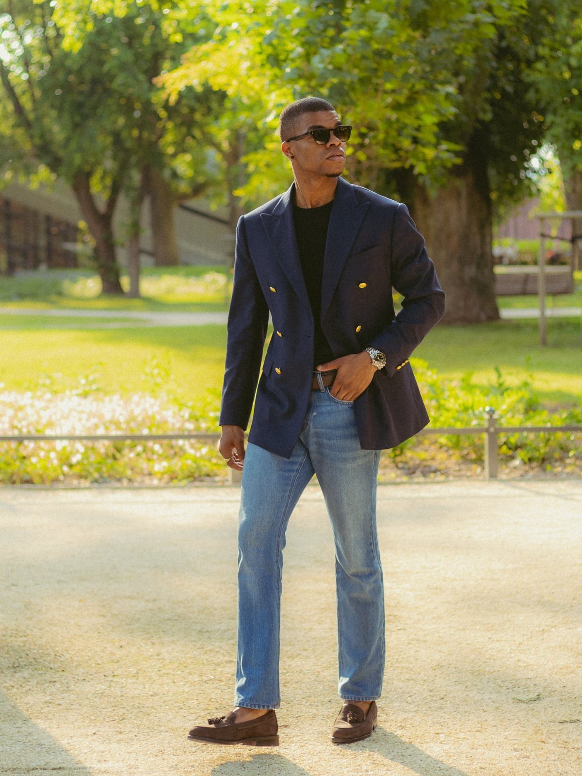 11 Blazer Outfits Casual Enough for Real Life 2 A man wearing a navy blazer, black shirt, and blue jeans standing in a park.