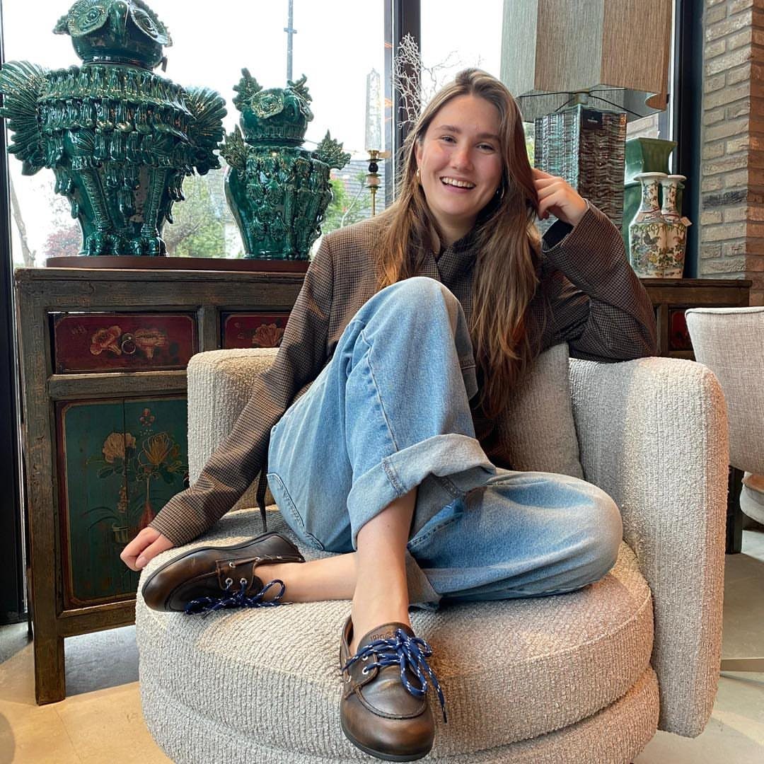 Smiling woman sitting in an armchair wearing a brown blazer, blue jeans, and brown boat shoes.
