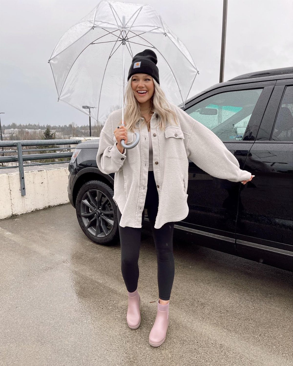 Smiling woman in a beige shirt, black leggings, and pink rain boots holding a clear umbrella.