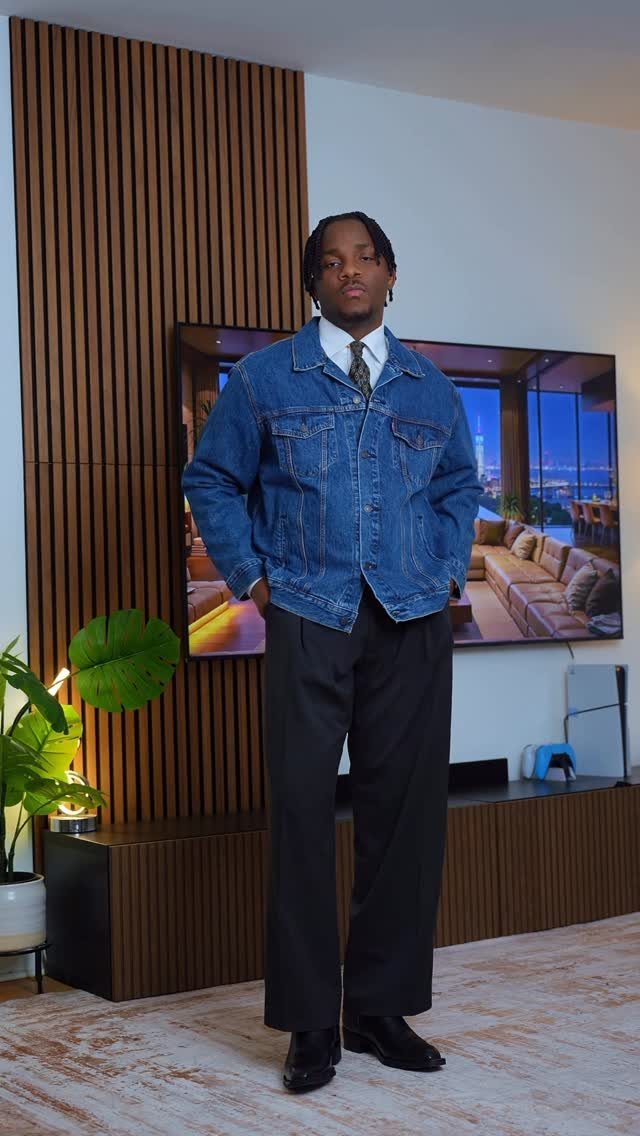 A person wearing an oversized denim jacket, white shirt, tie, and black wide-leg trousers.