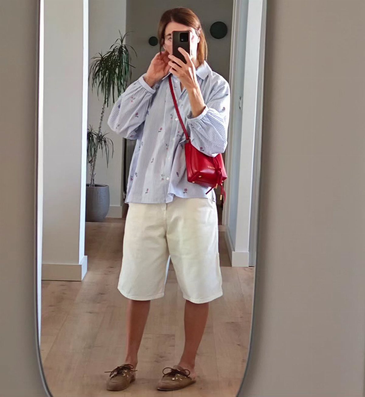 Woman in a striped shirt, white shorts, and tan boat shoes taking a mirror selfie.