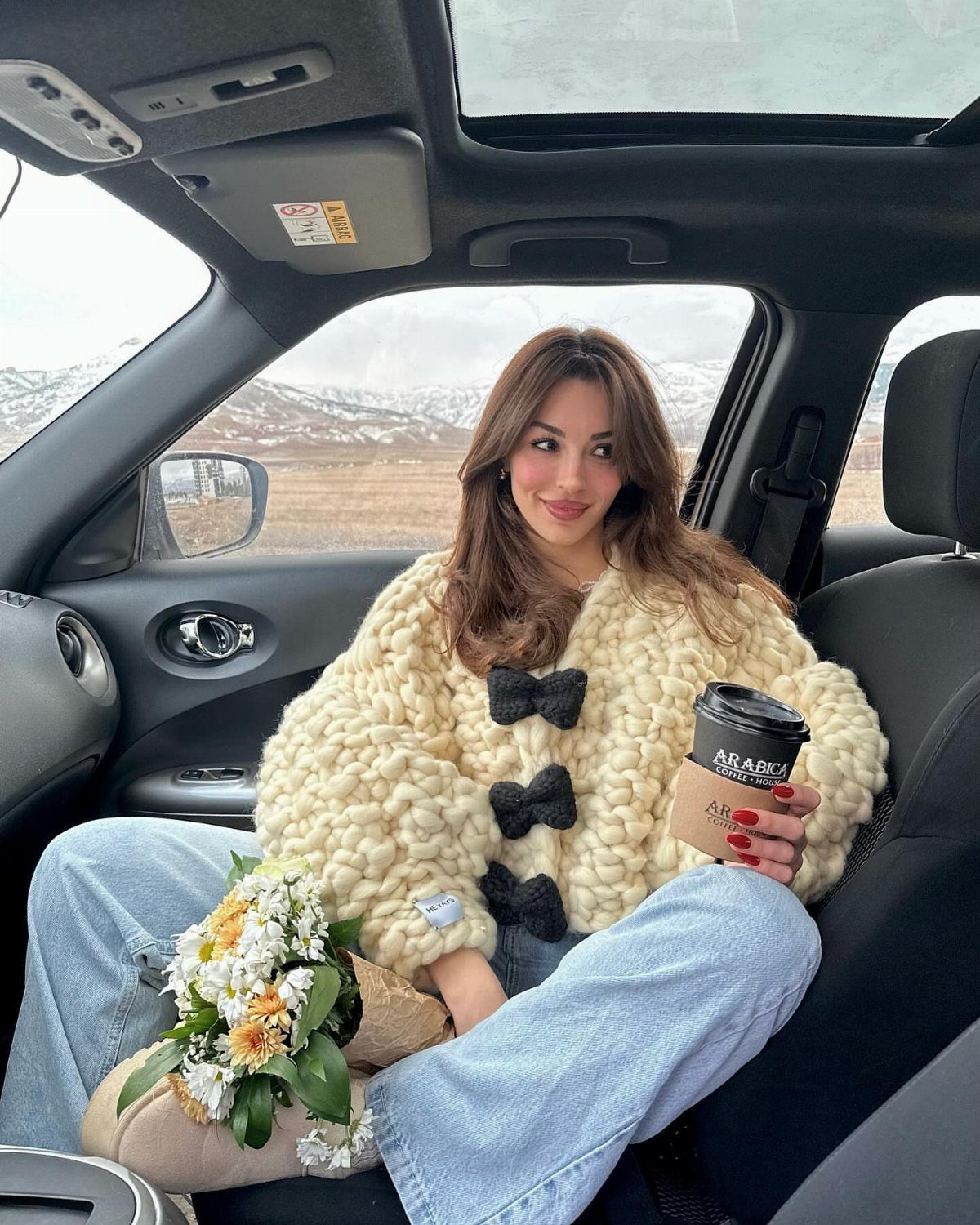 10 Winter Sweater Outfits: From Fair Isle to Full-On Neutrals 2 Woman in a car wearing a chunky cream knit cardigan with black bow details.