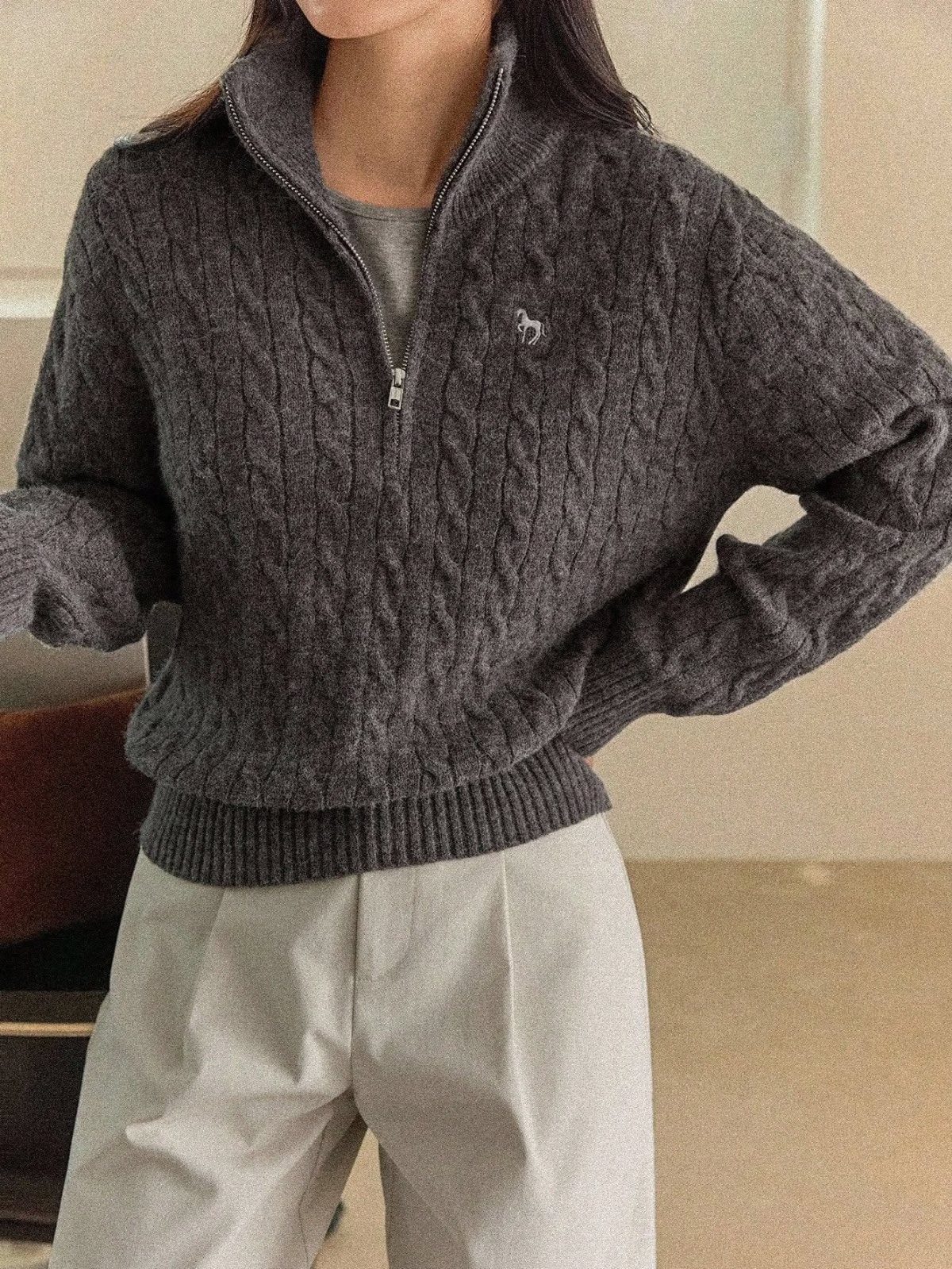 A person wearing a dark grey cable-knit quarter-zip sweater and light grey trousers.