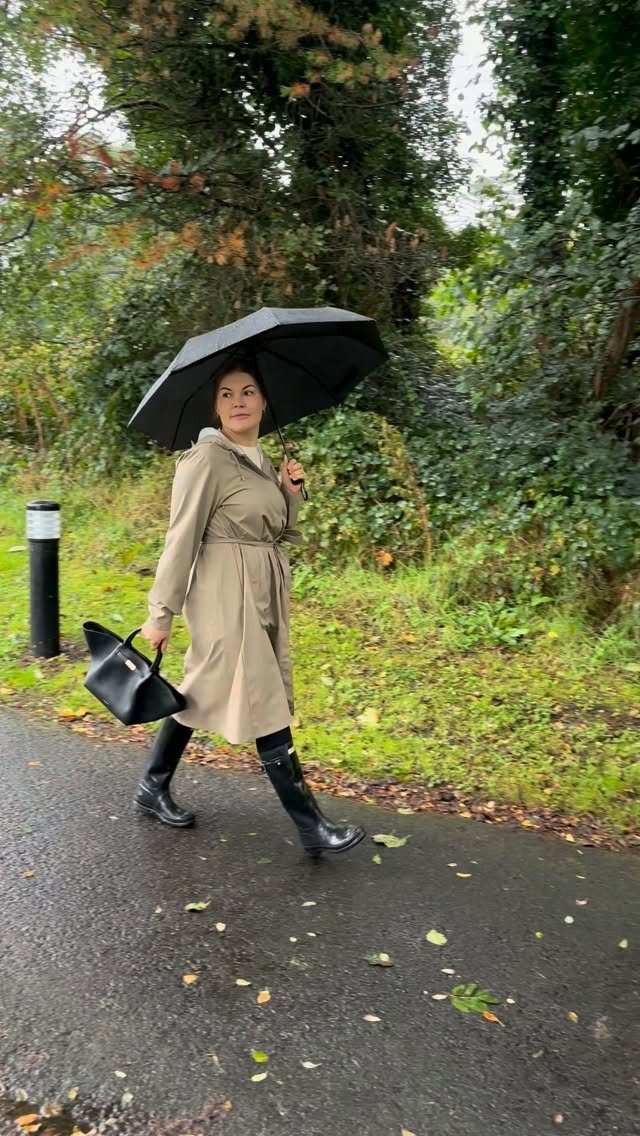 Woman in a beige trench coat and black boots walking with a black umbrella.