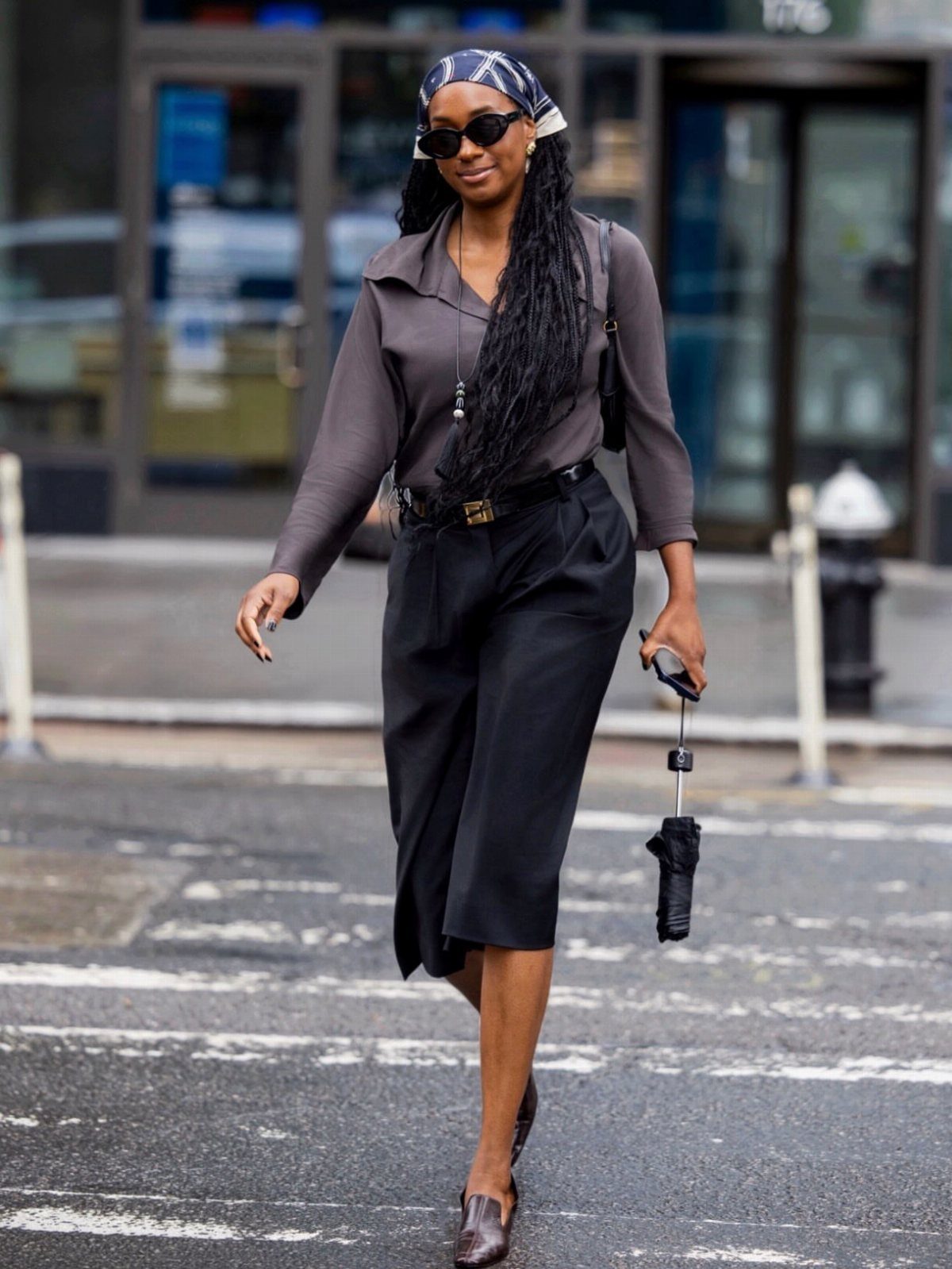 A woman walking on a city street wearing a grey blouse and black cropped trousers.