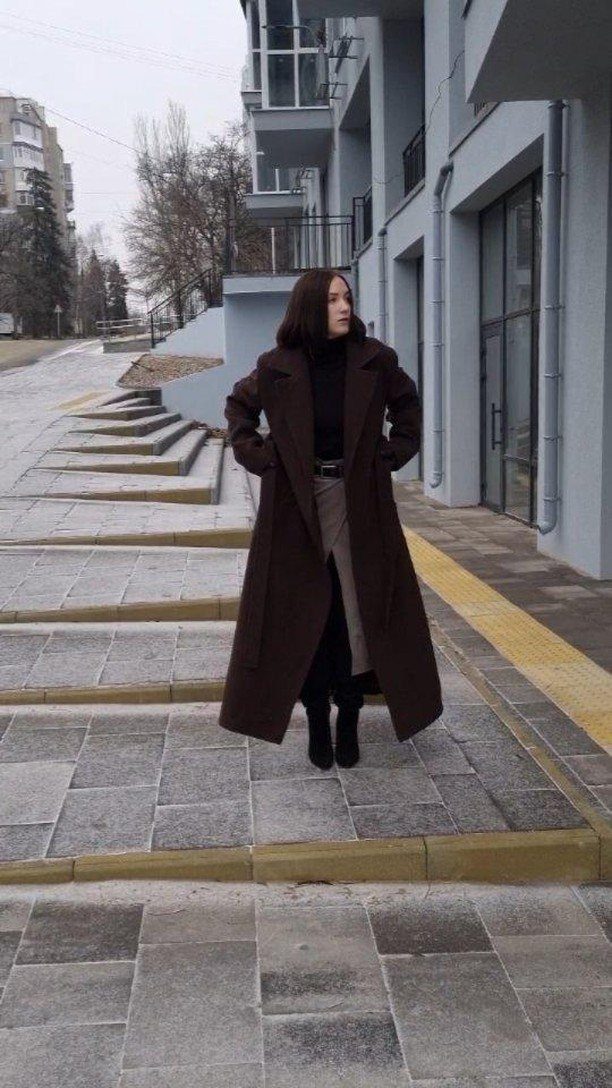 A woman wearing a long dark brown wool coat standing on a stone walkway outdoors.