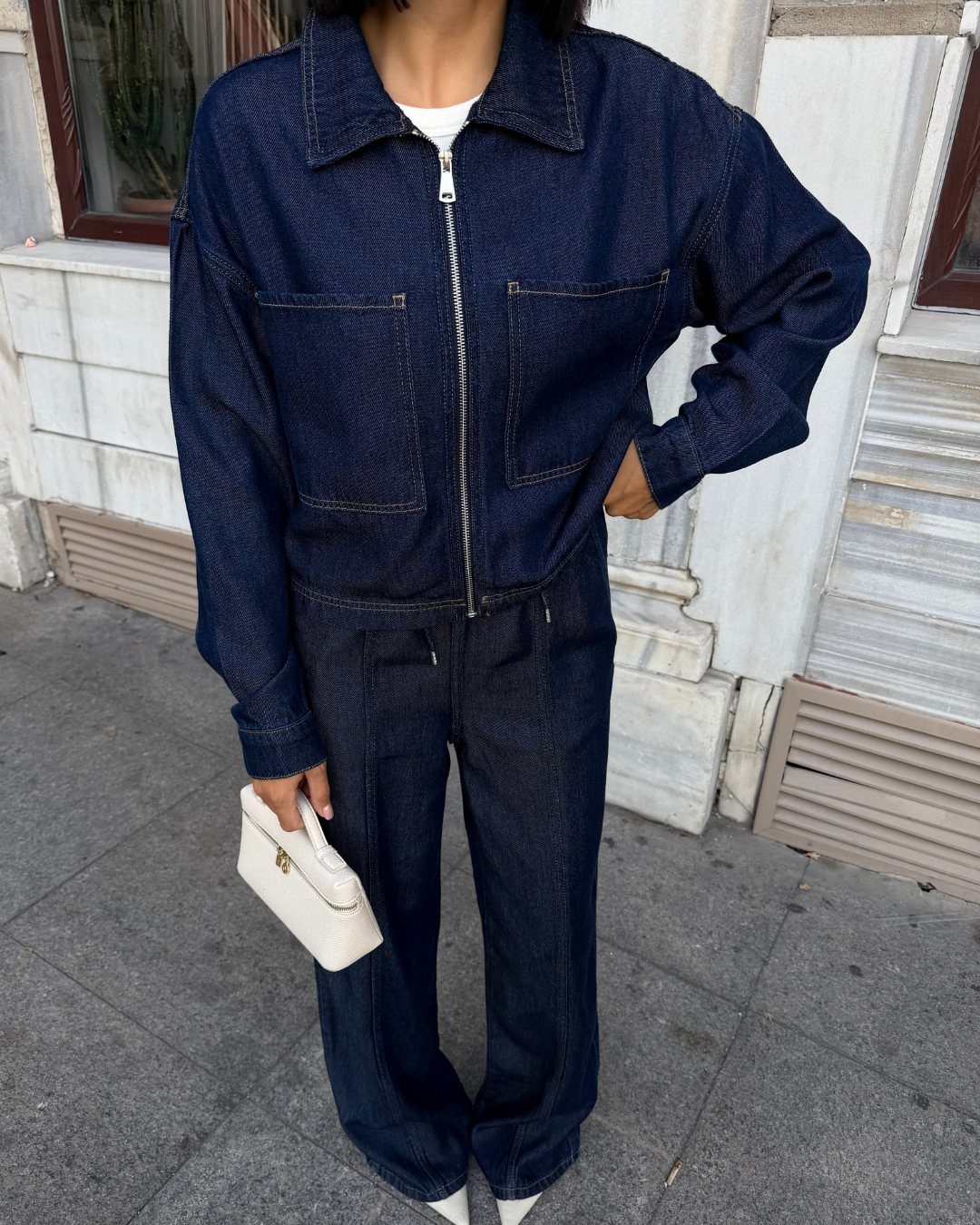 A person wearing an oversized dark denim jacket and matching denim pants holding a white bag.