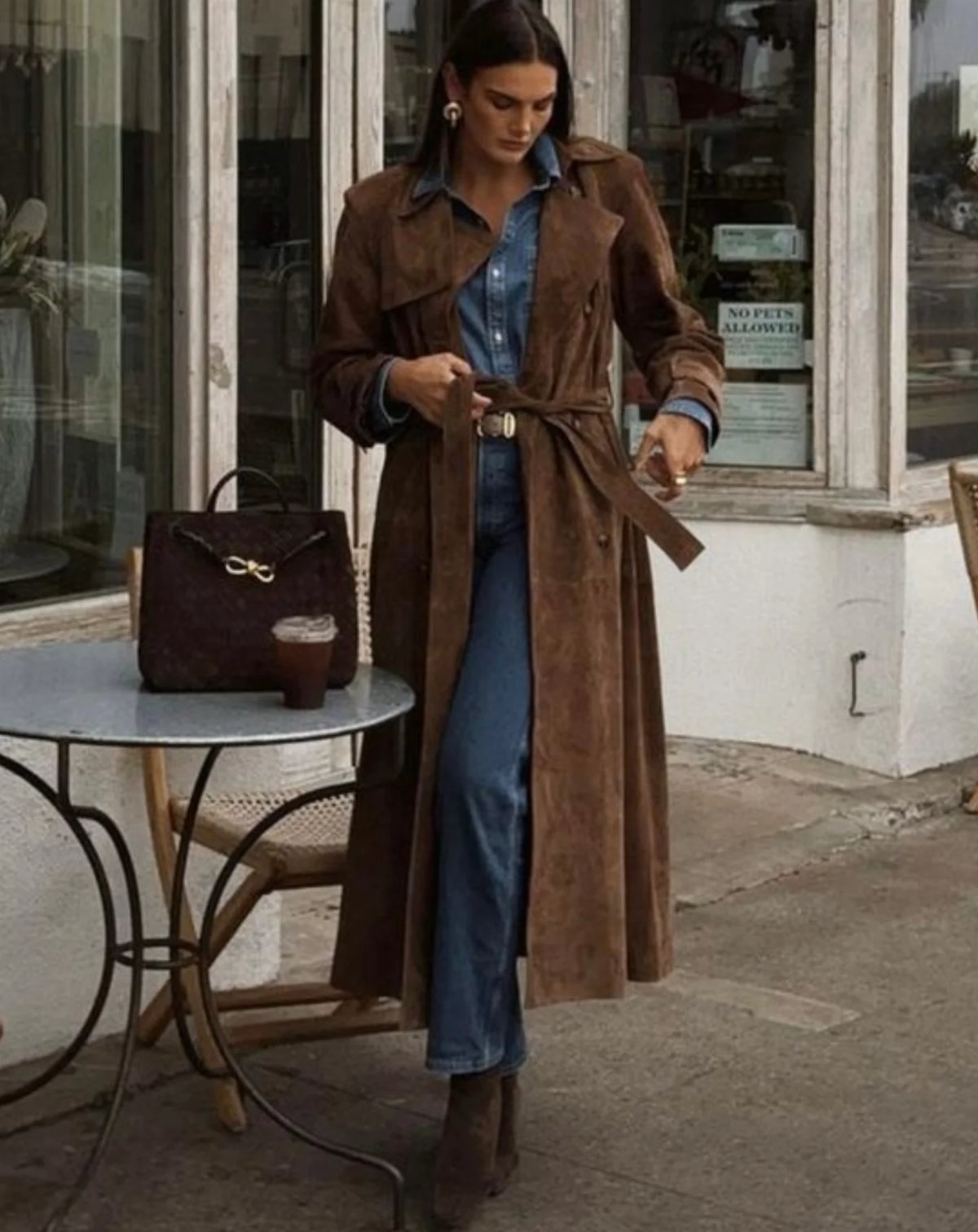 A woman wearing a long brown suede duster coat and blue denim jumpsuit outdoors.