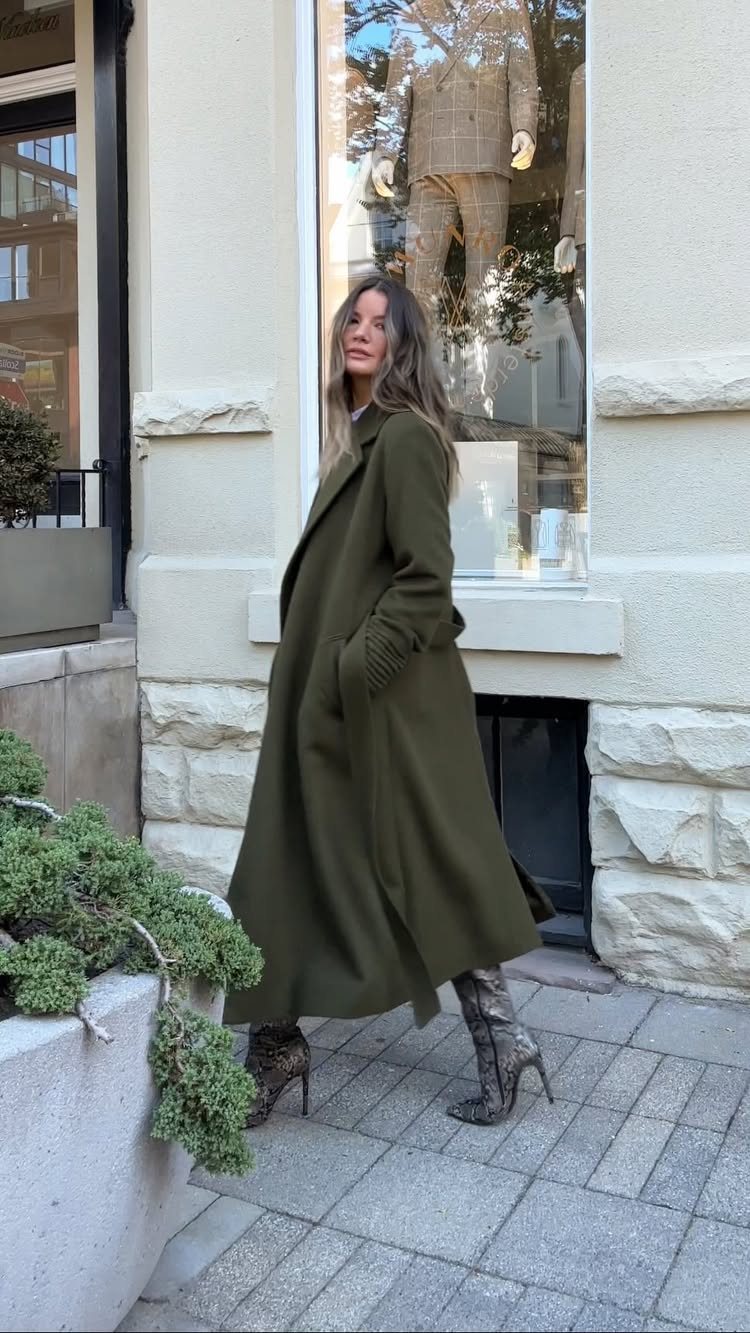A woman posing in a long olive green duster coat and patterned high-heeled boots.