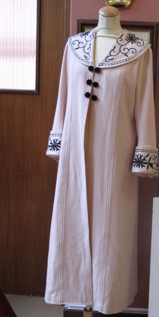 A long light pink coat with black embroidery on the collar and cuffs displayed on a mannequin.