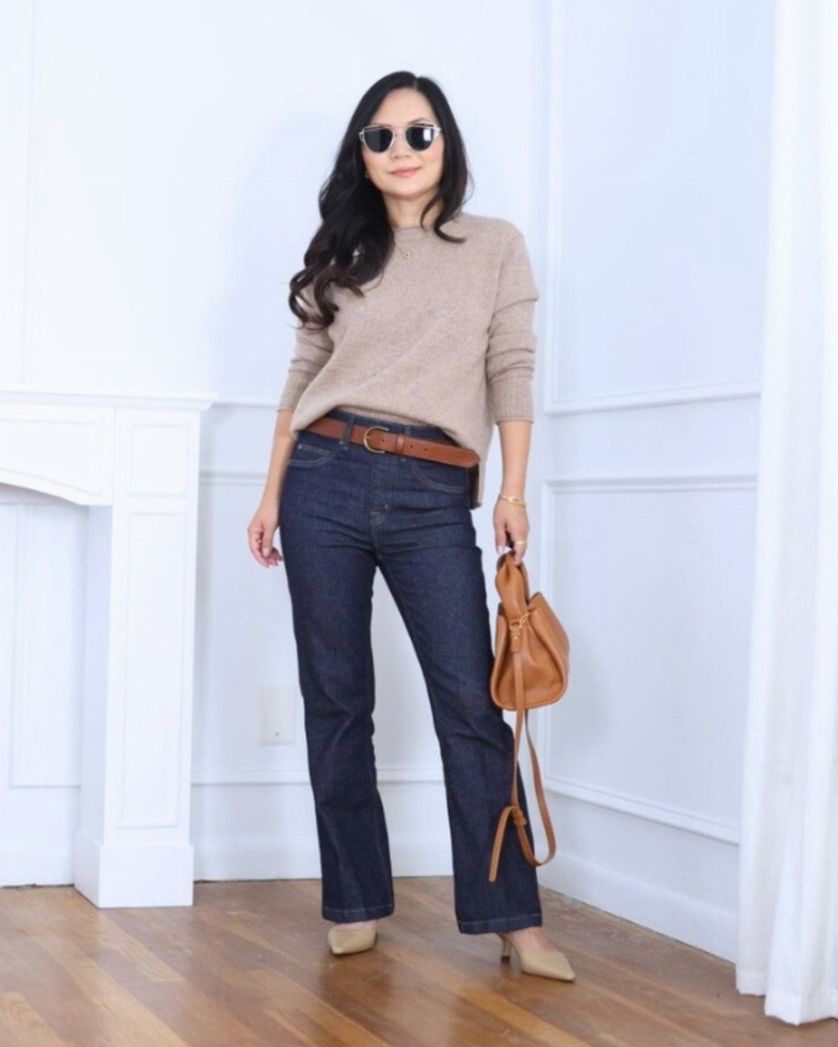 Woman wearing a beige sweater, dark blue flared jeans, and beige heels standing in a white room.