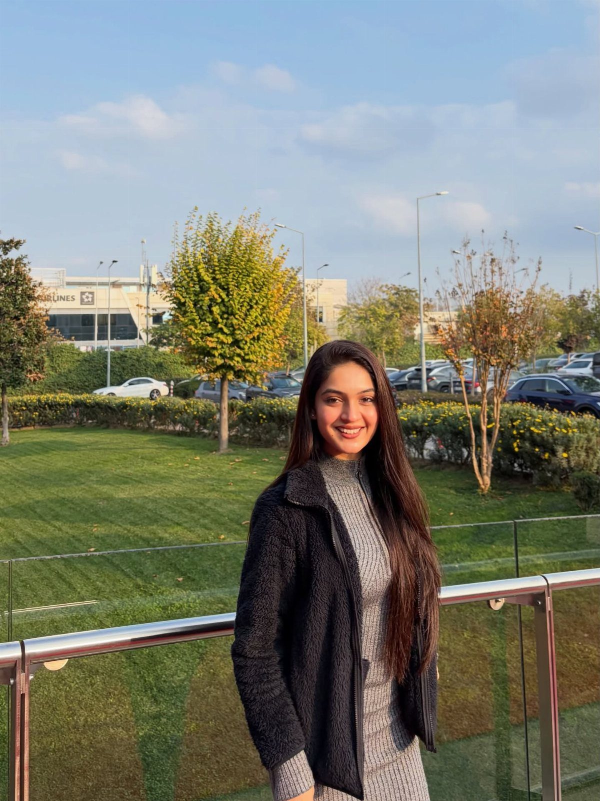 A woman wearing a dark fleece jacket over a grey ribbed knit dress smiling outdoors.