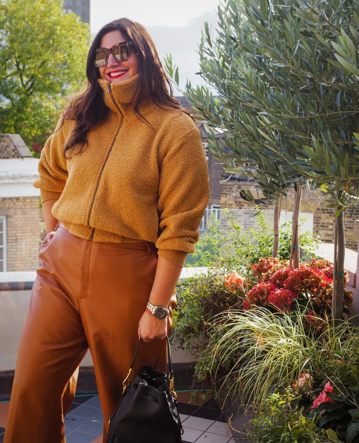 Woman smiling in a mustard fleece pullover and brown wide-leg tailored trousers outdoors.