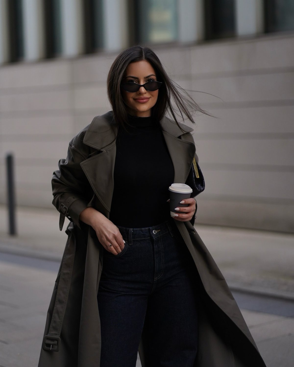 Woman in a dark green trench coat, black turtleneck, and sunglasses walking on a city street.