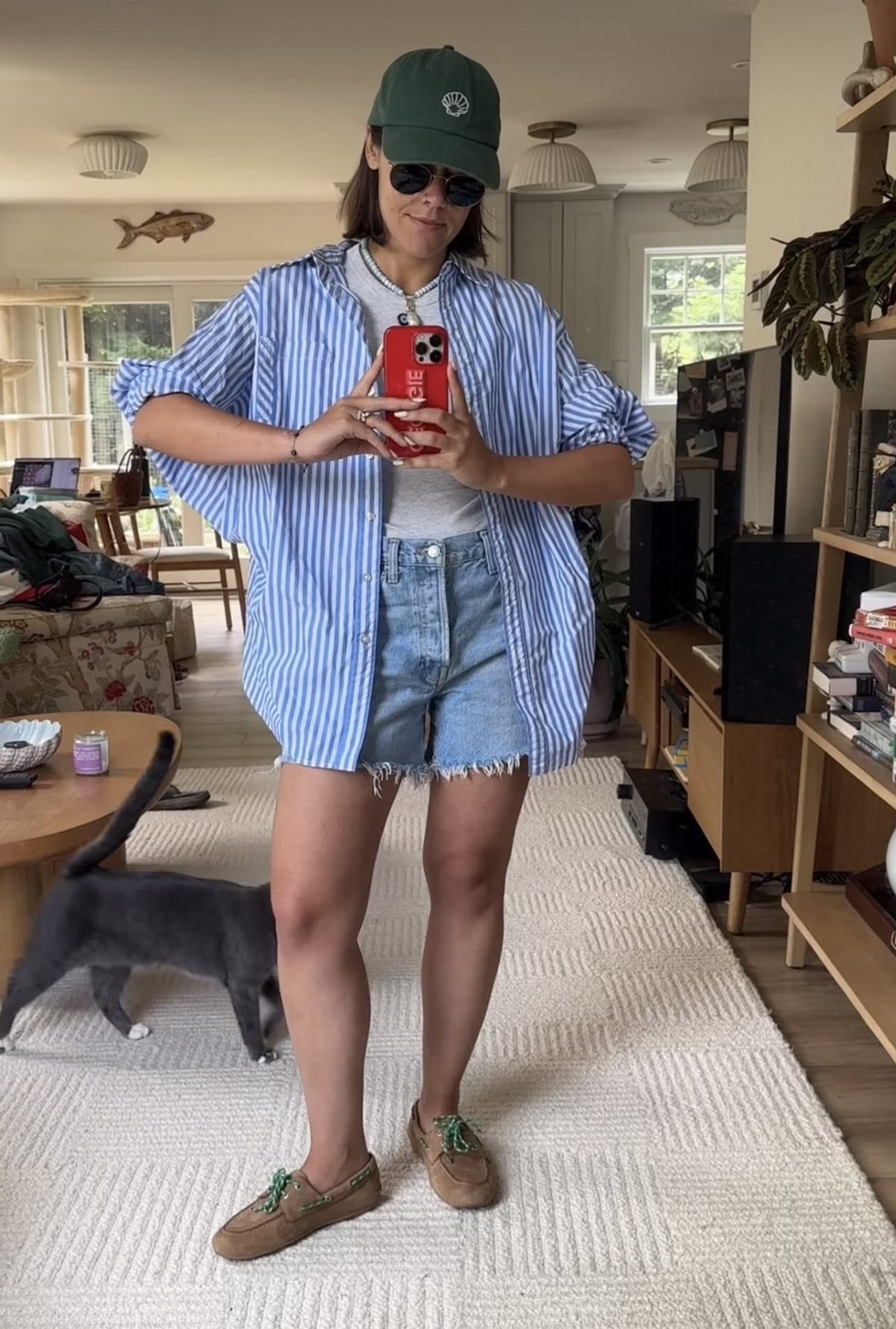 Woman in striped button-up shirt and denim shorts posing for a mirror selfie.
