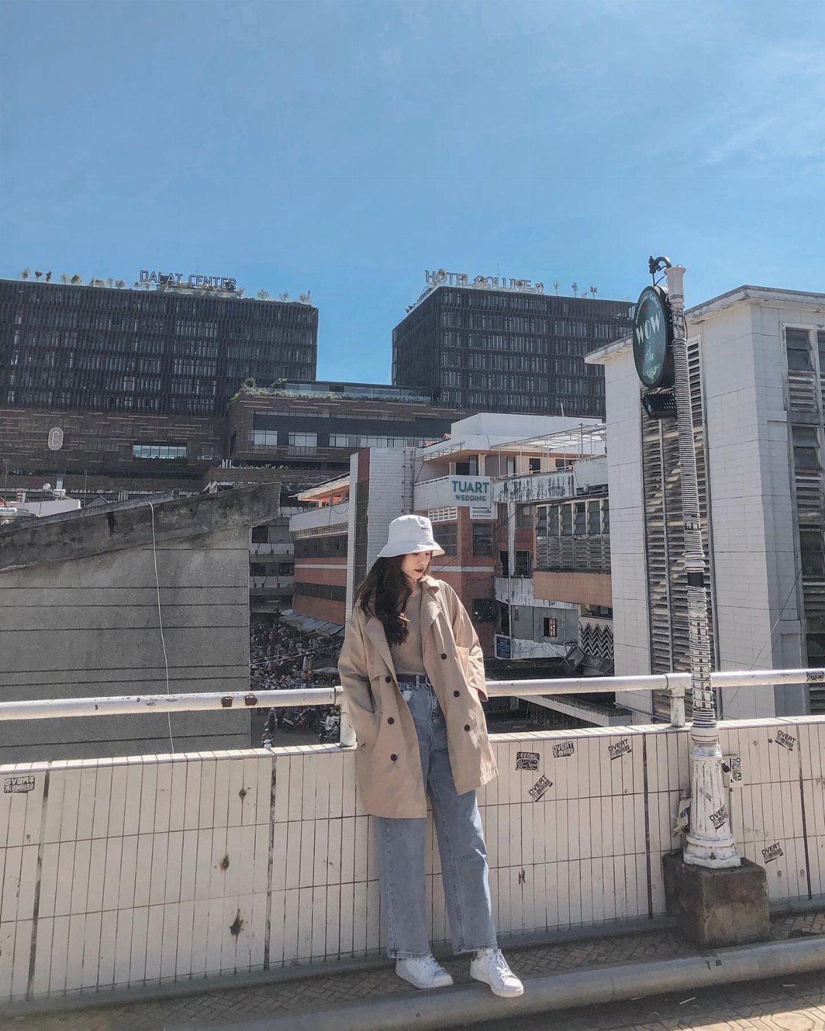 Woman in a long beige trench coat, light blue jeans, and white bucket hat standing outdoors.