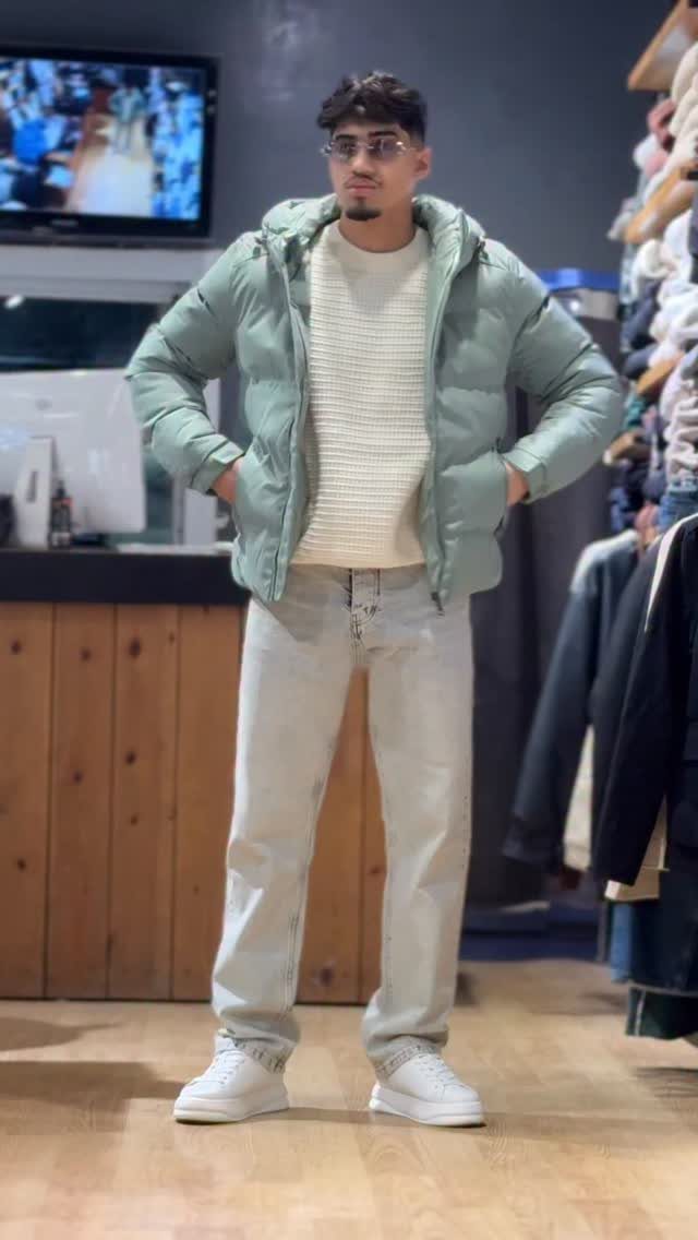 Man wearing a light green puffer jacket, cream sweater, and light wash jeans.