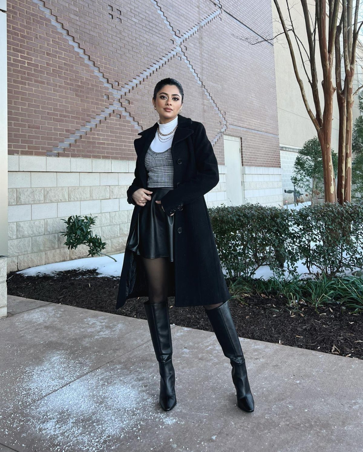 15 Long Boots Outfit Ideas for Cold Months Ahead 9 Woman in a long black coat, mini skirt, and black knee-high leather boots standing outdoors.