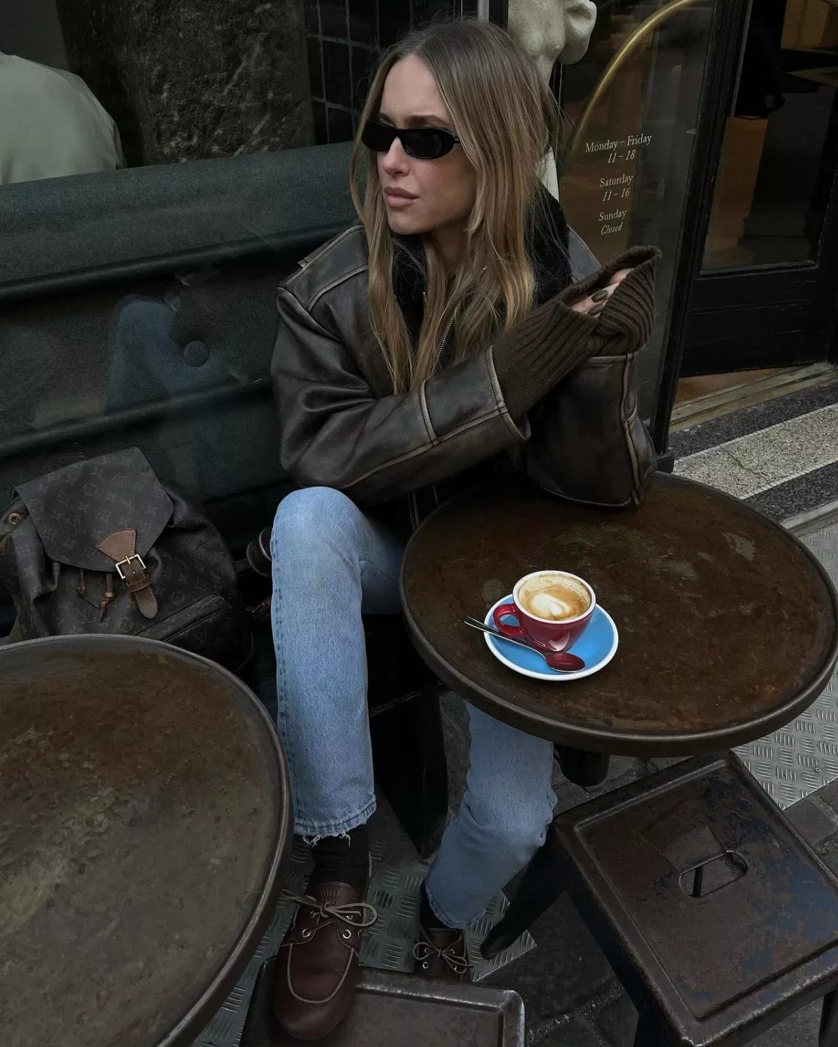 Woman sitting at a cafe wearing a brown leather jacket, light blue jeans, and brown boat shoes.