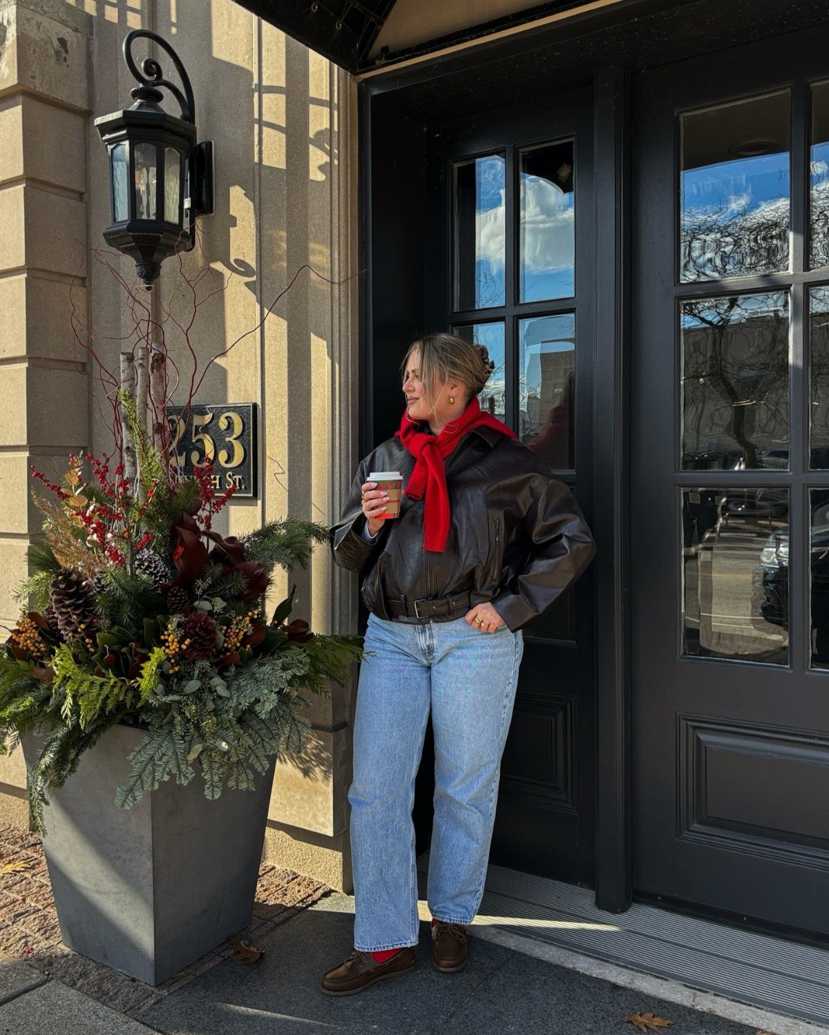 Woman wearing a black leather jacket, red scarf, and light blue jeans standing outdoors.