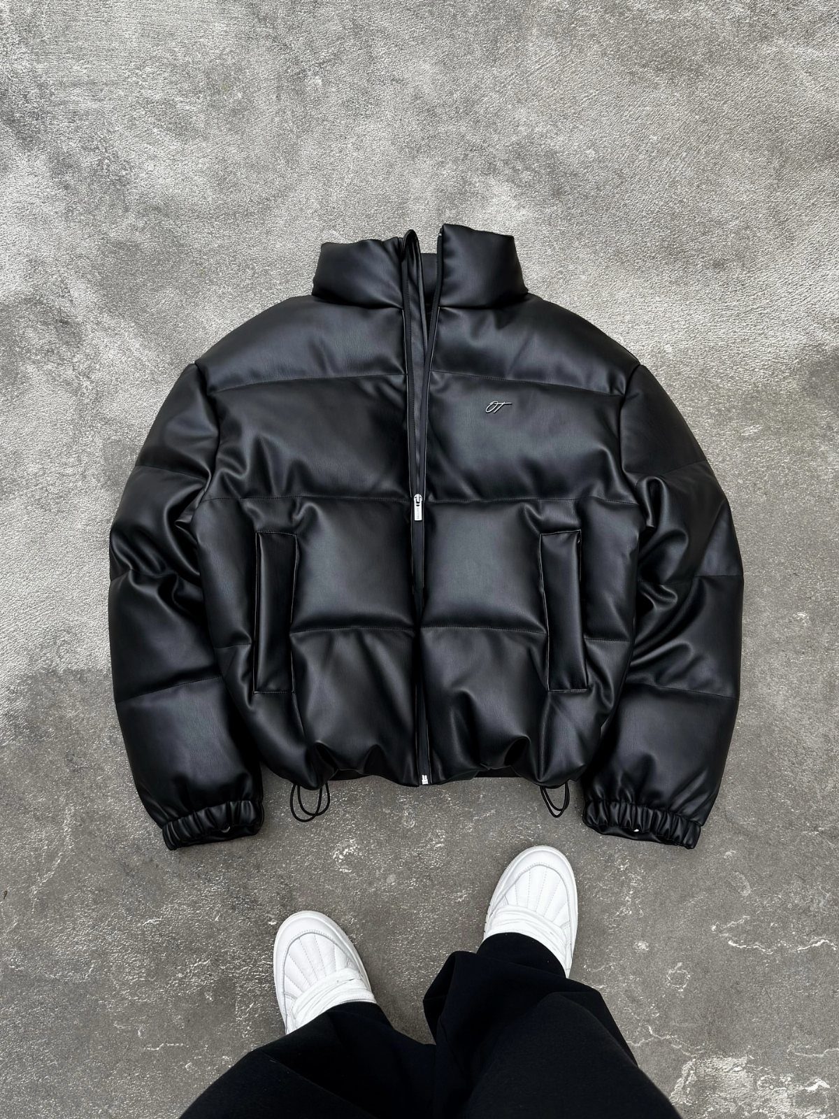 Black leather puffer jacket laid on grey pavement with black trousers and white sneakers below.