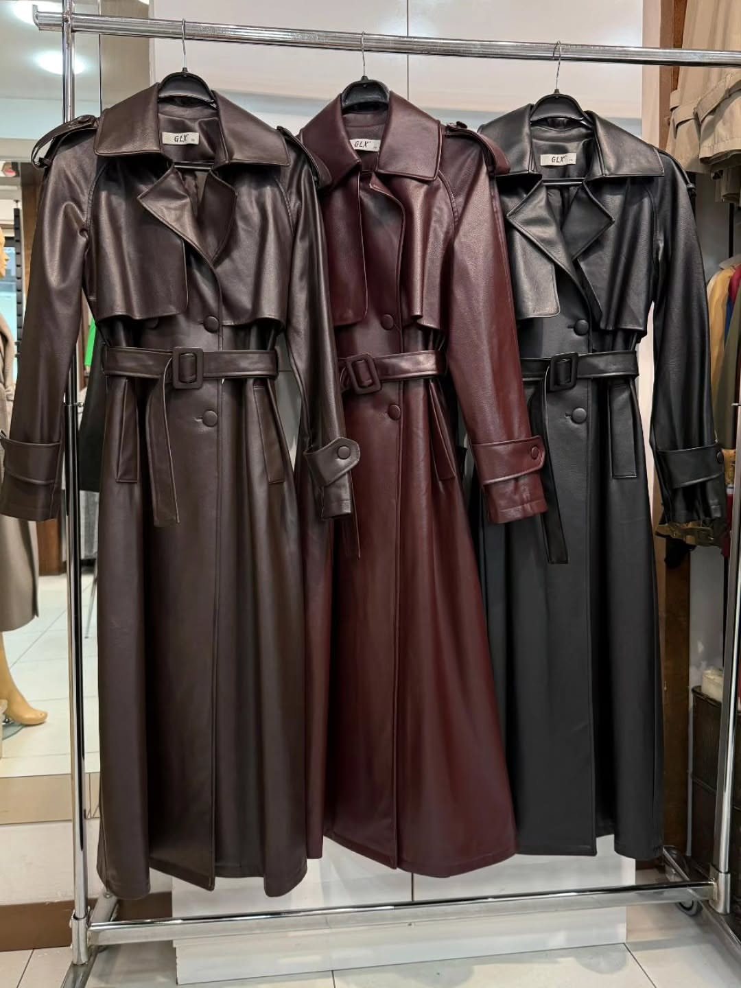 Three long leather trench coats in brown, burgundy, and black hanging on a metal clothing rack.