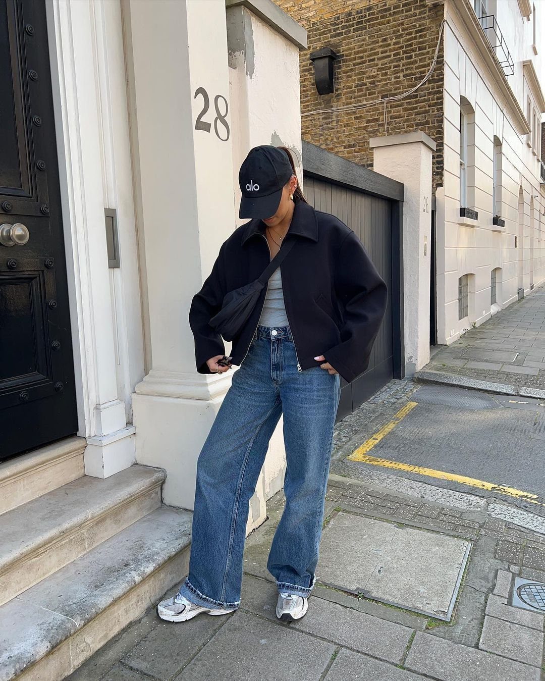 Person wearing a black jacket, blue jeans, and silver trainers standing on a city sidewalk.