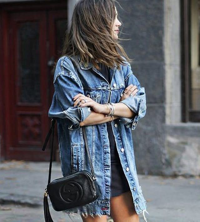 Woman wearing an oversized long denim jacket and a black crossbody bag while standing outdoors.