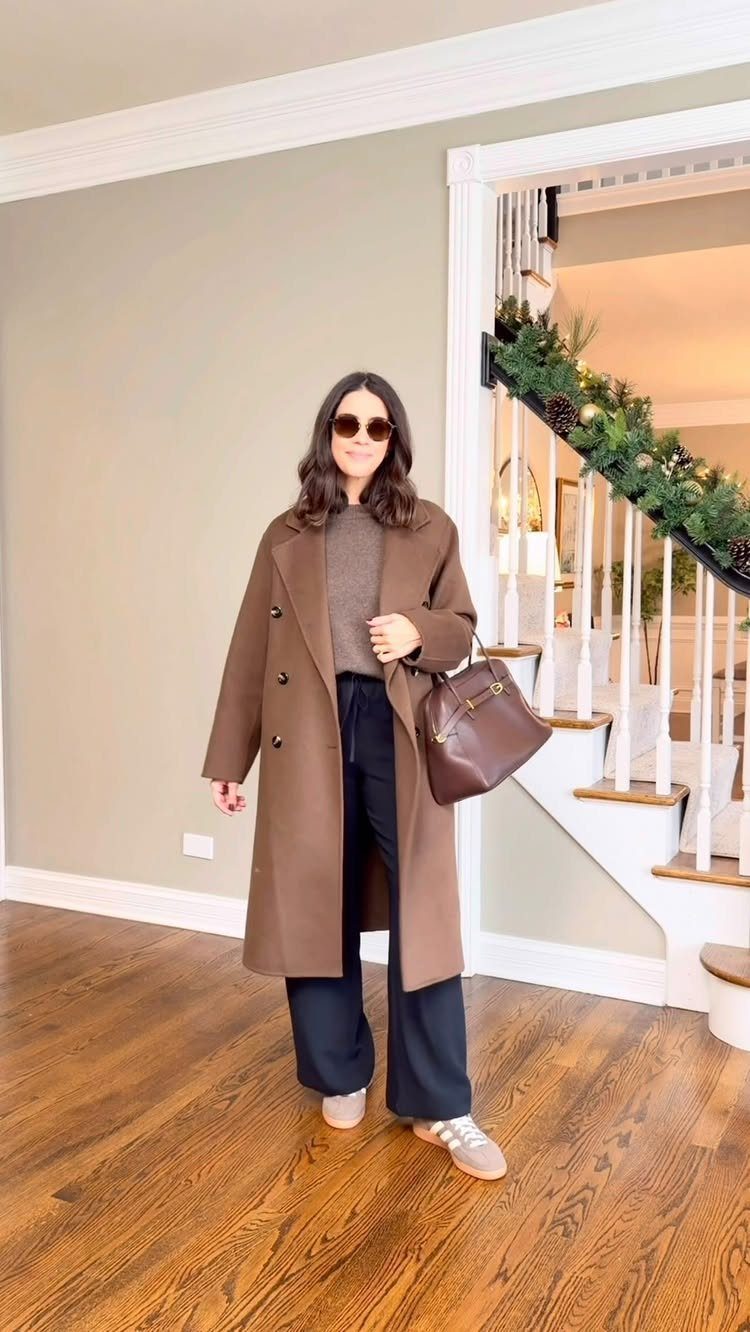 Winter Capsule Wardrobe for Women: 12 Ideas That Actually Work 8 Woman wearing a long brown coat, grey turtleneck, black trousers, and beige sneakers indoors.