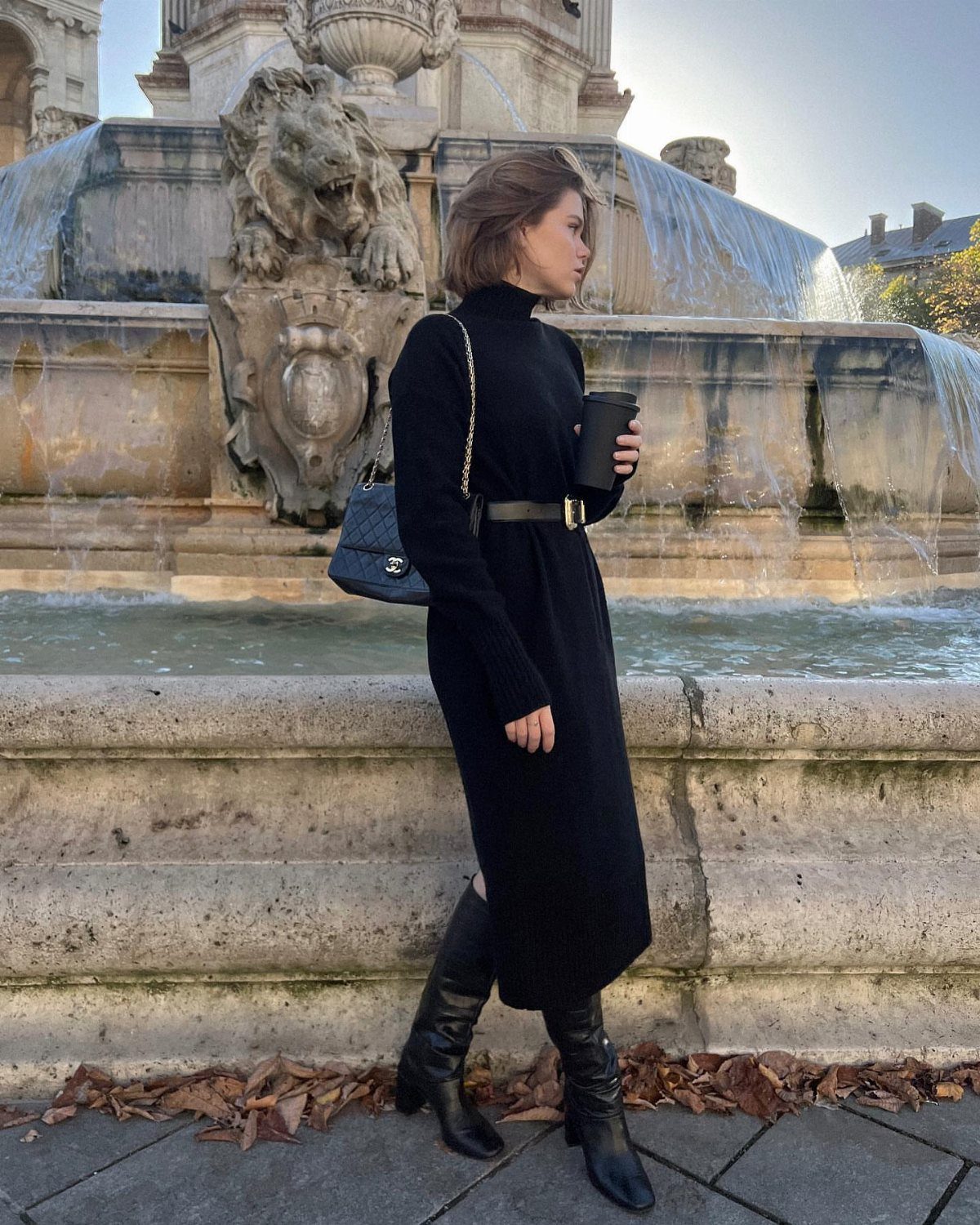15 Long Boots Outfit Ideas for Cold Months Ahead 4 Woman in a black turtleneck midi dress and black leather knee-high boots standing by a fountain.