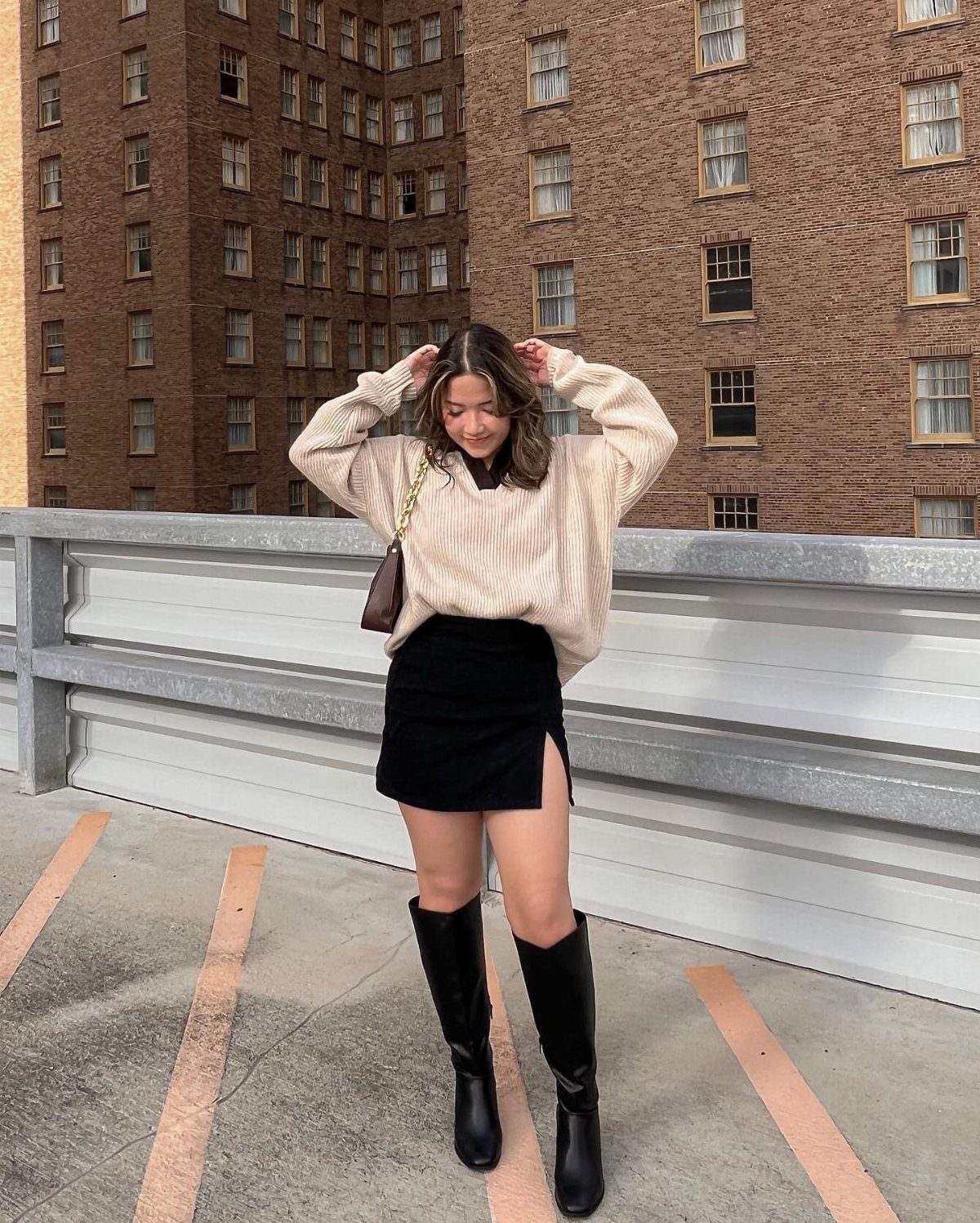 15 Long Boots Outfit Ideas for Cold Months Ahead 2 Woman wearing a cream sweater, black mini skirt, and tall black boots on a rooftop.