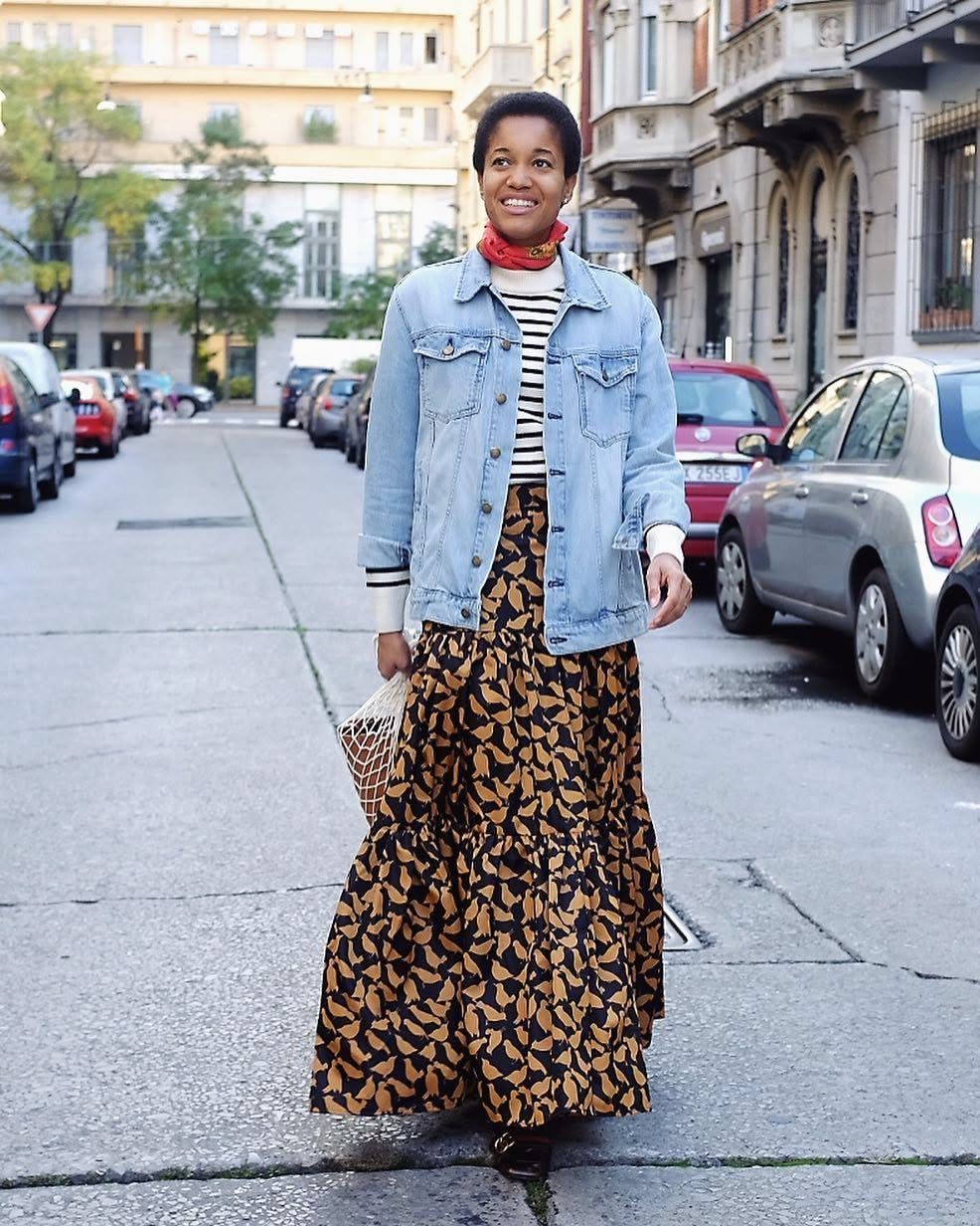 Long Denim Jacket Outfit: 8 Looks to Wear Now 7 Woman walking in a city wearing a denim jacket, striped top, and patterned maxi skirt.
