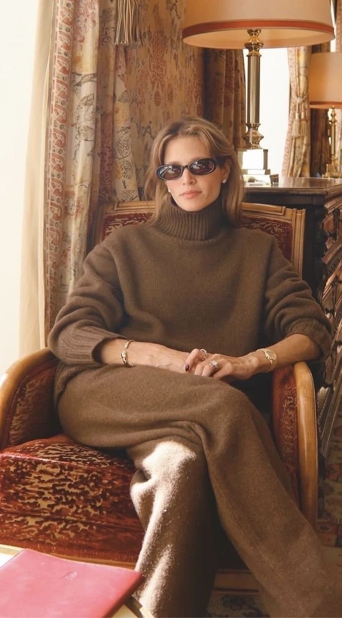A woman in a brown turtleneck sweater and trousers sitting in an armchair wearing sunglasses.