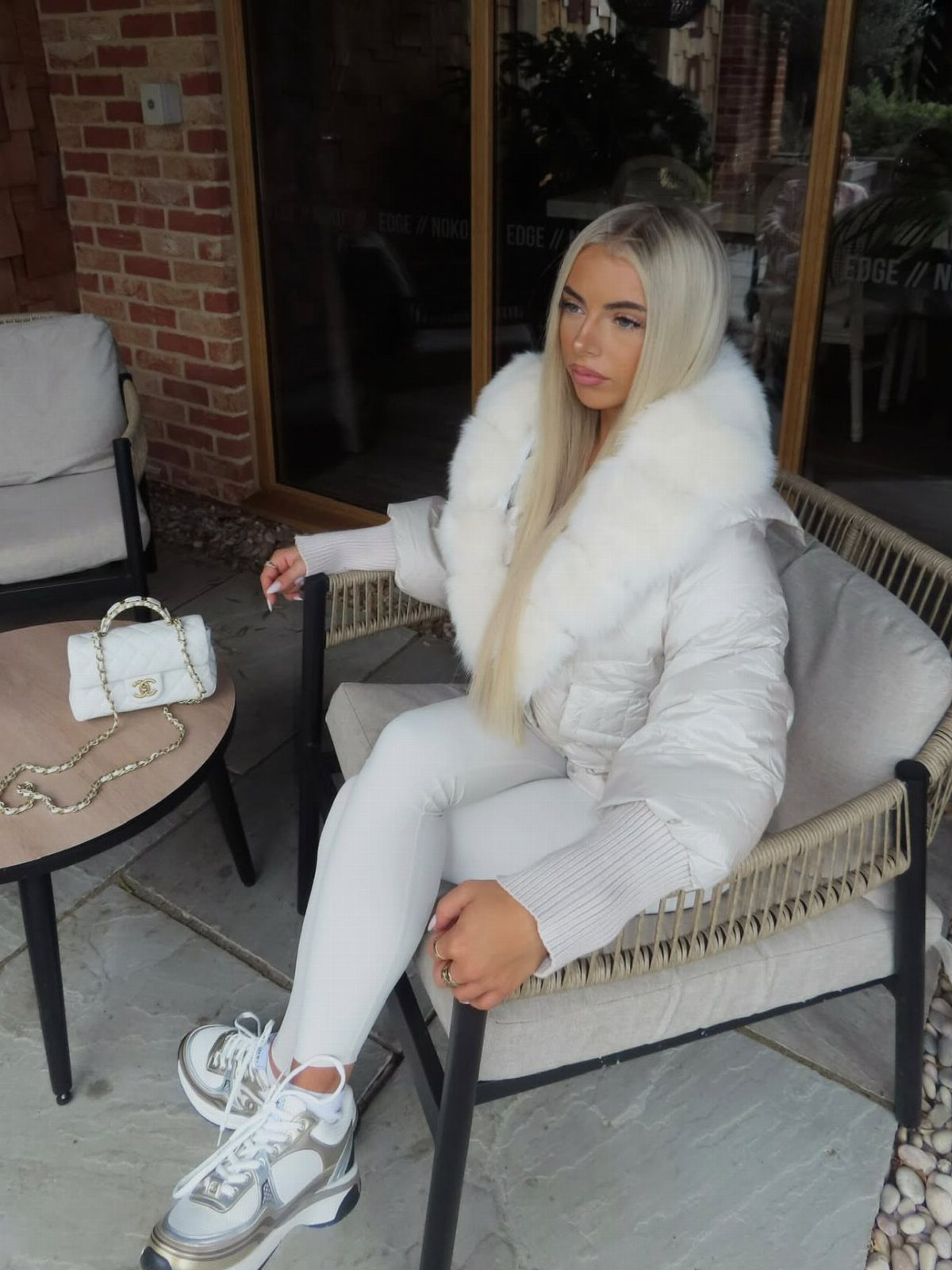 Blonde woman sitting in a chair wearing a white puffer jacket, white leggings, and silver sneakers.