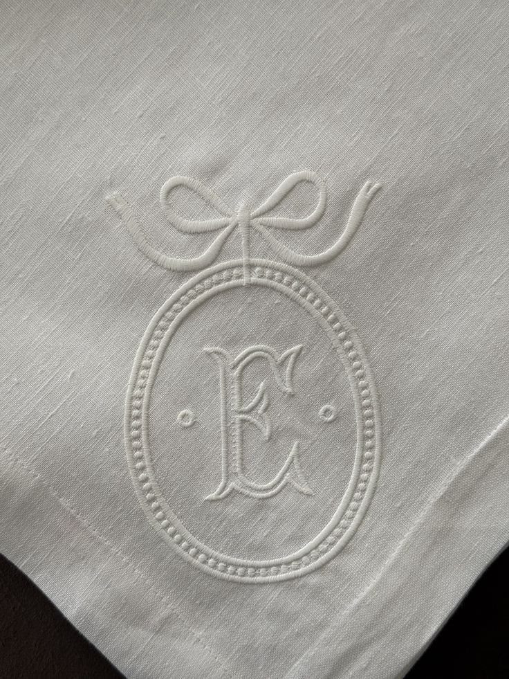 White embroidered monogram of the letter E inside an oval frame with a bow on linen.