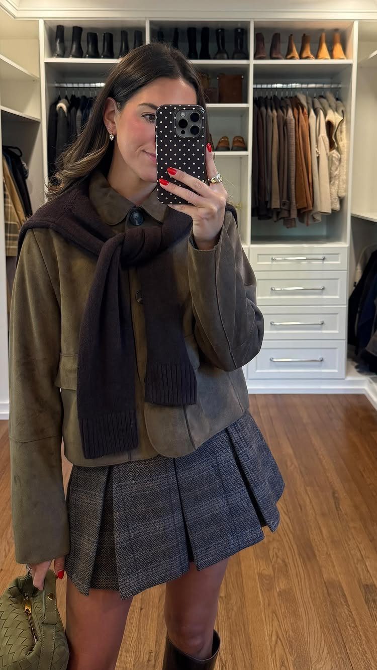 Woman in olive jacket and plaid skirt posing for a mirror selfie in a closet.