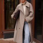 Blonde woman wearing a long beige wool coat and light blue flared jeans outdoors.