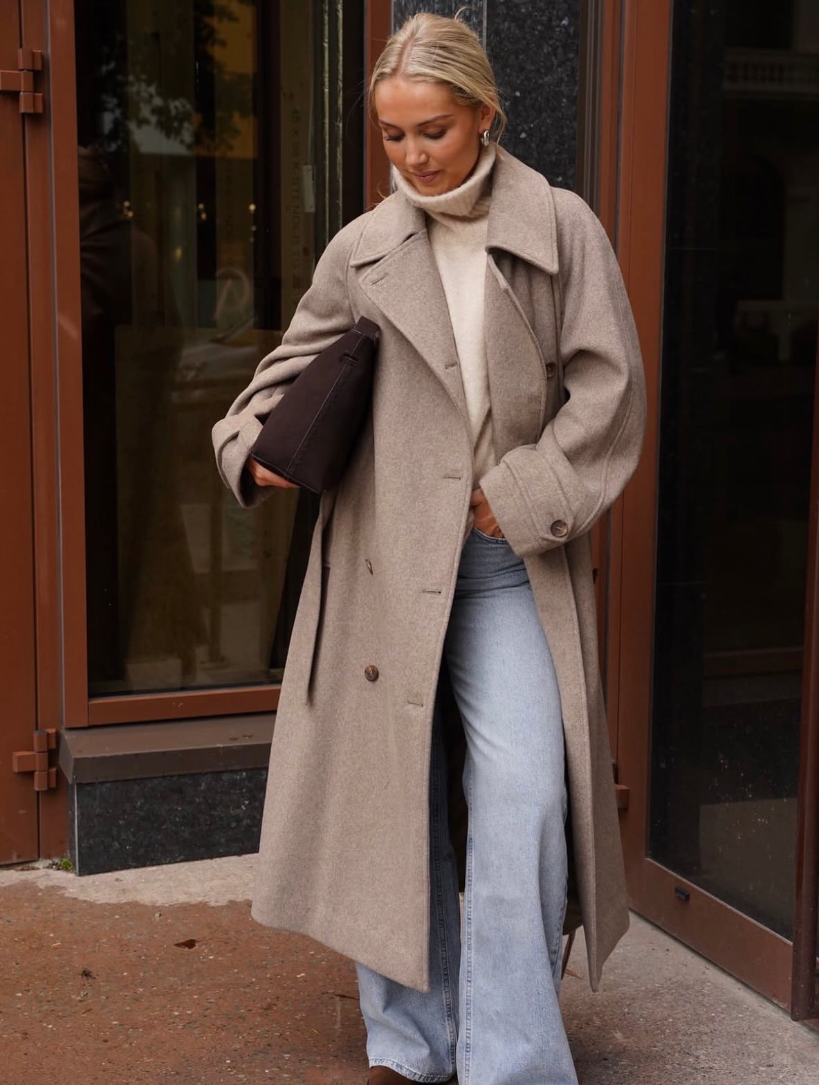Blonde woman wearing a long beige wool coat and light blue flared jeans outdoors.