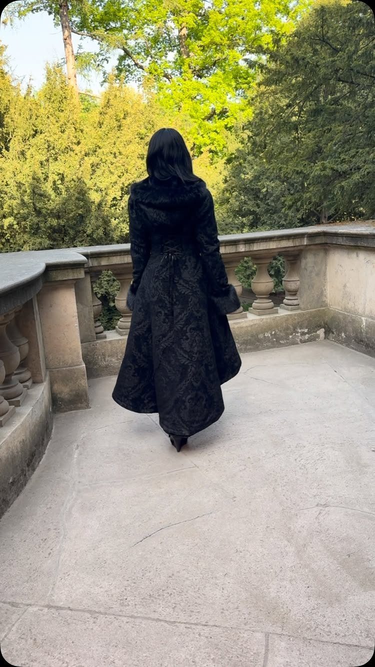 Opera Coat Women: 6 Vintage Styles Worth Wearing 2 Woman in a long black textured opera coat standing on a stone balcony with trees.