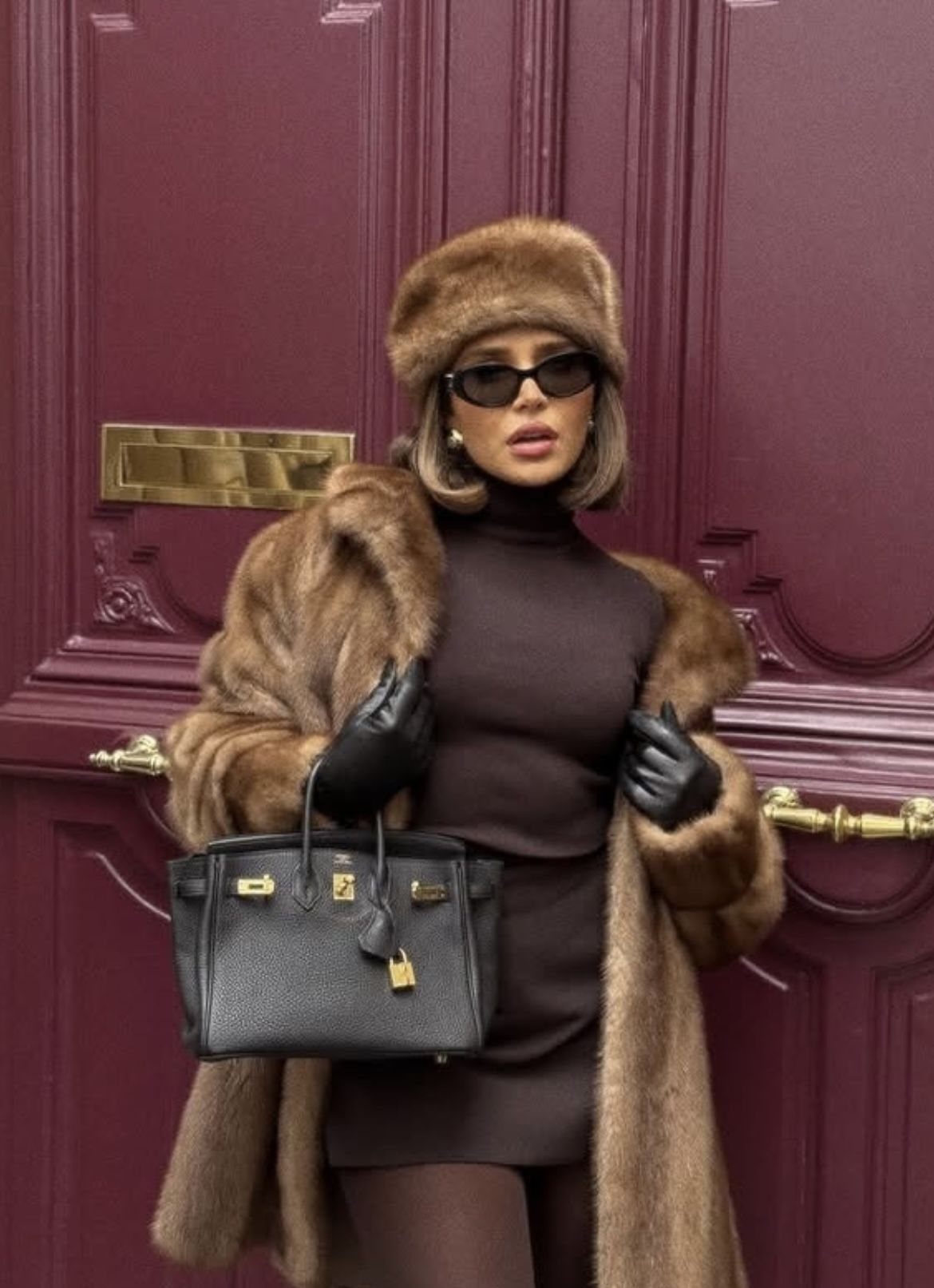 Woman in a brown fur coat and hat posing with a black handbag.