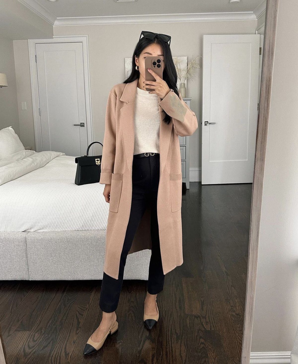 Woman in a long beige coat, dark trousers, and pointed-toe flats posing for a mirror selfie.