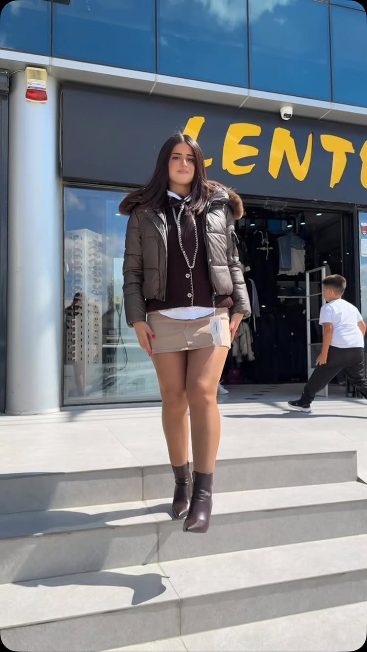 Woman in a brown puffer jacket, dark vest, and tan mini skirt walking down stairs.