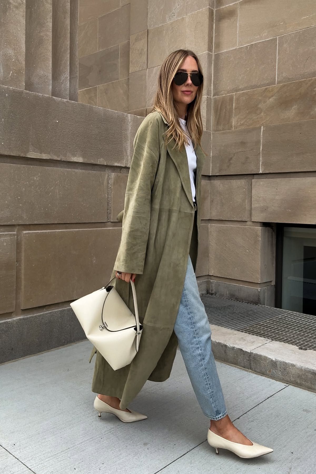 Woman walking in an olive green trench coat, white top, and light blue jeans.