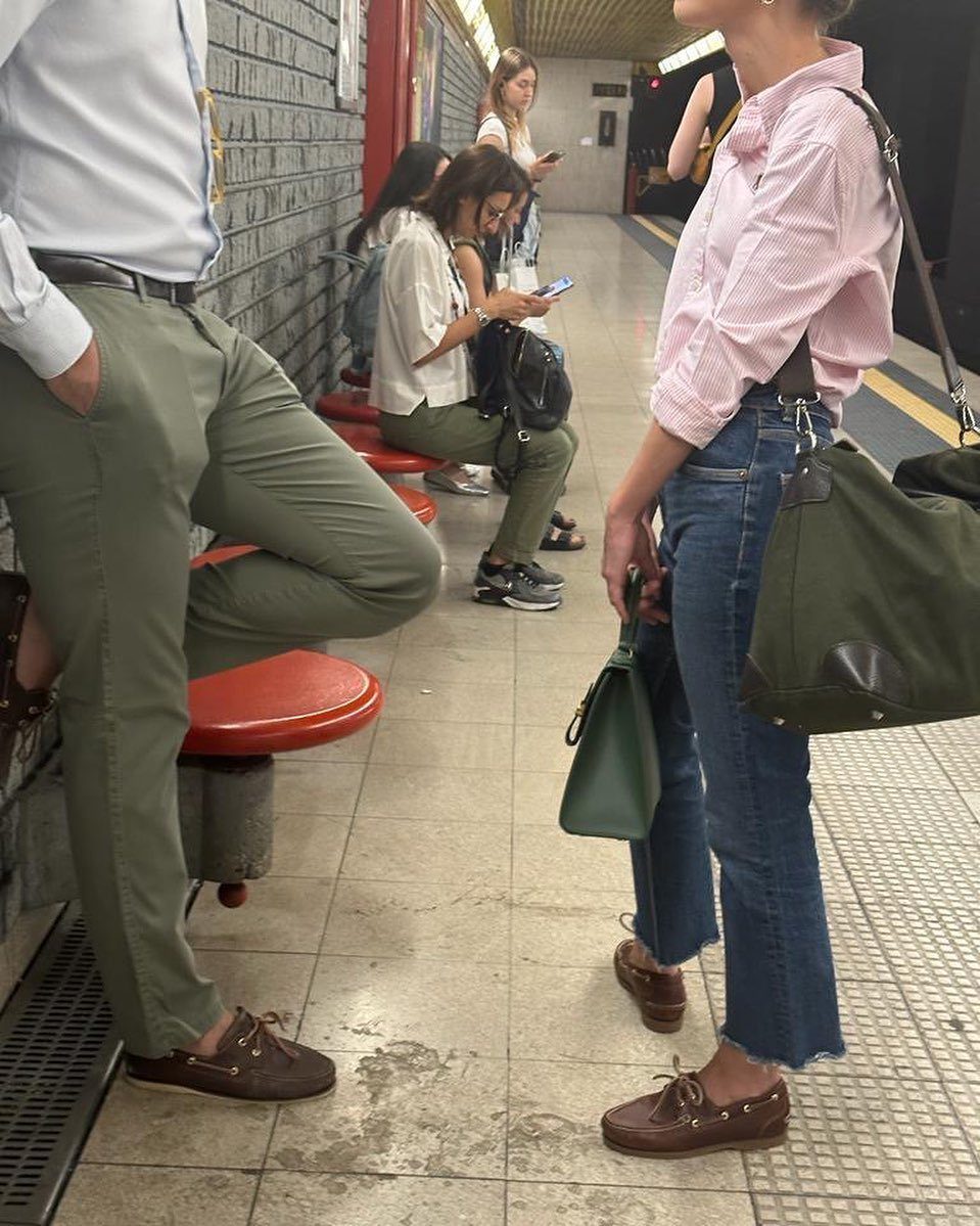 A woman in a pink striped shirt, blue jeans, and brown boat shoes standing in a subway.