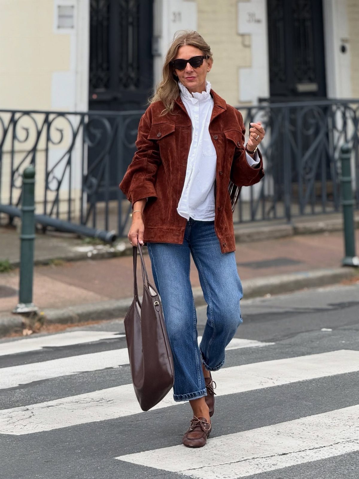 Woman walking in a crosswalk wearing blue jeans, a white shirt, brown suede jacket, and boat shoes.